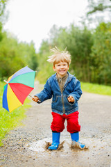 Child jumps on puddles in boots. Baby in the rain. A boy with a rainbow umbrella is walking outside. Autumn, vacation