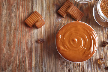 Sweet caramel candies and bowl with sauce on wooden table
