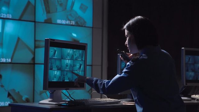 Single female security person monitoring closed circuit surveillance from multiple cameras in various rooms of a building informs on the radio to check the person who moves inside the room.
