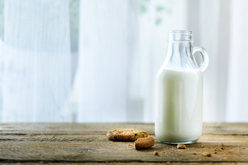 Chocolate chip cookies, bottle and glass of milk on wooden table near window, white background. Sunny morning, copy space