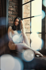 Woman sitting near window, light glow blur. Happy smiling young lady in white long sweater and Cashmere socks