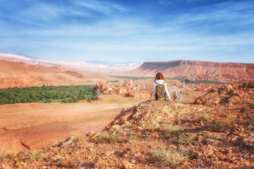 Young woman looking at fortified city. Glaoui Kasbah of Telouet kasbah or ksar in Morocco view from above. Traveler girl sits on a rock looks up at the valley of Ounilla - obrazy, fototapety, plakaty
