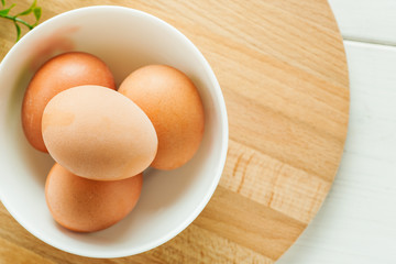Chicken eggs in a white ceramic bowl on a wooden round board with greenery, top view