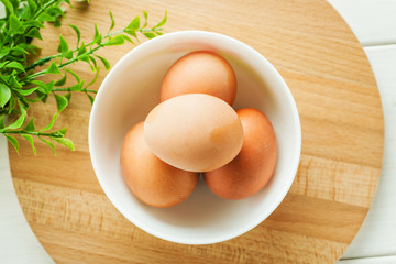 Chicken eggs in a white ceramic bowl on a wooden round board with greenery, top view