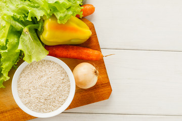 Fresh vegetables with rice in a white ceramic bowl on a wooden cutting board on a white table, top view