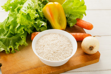 Fresh vegetables with rice in a white ceramic bowl on a wooden cutting board on a white table