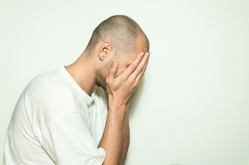 Young depressed man suffering from anxiety and feeling miserable cover his face with his hands and leaning on the white wall
