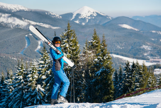 Female skier standing on top of a hill wearing blue ski suit mask and black helmet, holding skis on the shoulder, smiling to the camera. Winter snow mountains nature recreation resort on background