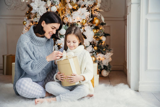 Portrait of pretty girl and beautiful female mother sit together on warm carpet, hold present box, enjoy decorated New Year tree. Family spend time together. Celebration and holidays concept