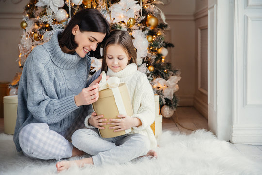 Indoor shot of adorable girl and her mother sit crossed legs, open wrapped present box, have intrigue what is there, sit against adorned New Year or Christmas tree, have sincere gentle smiles on faces
