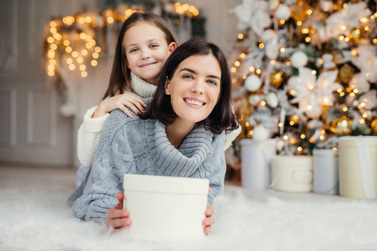 Indoor shot of mother and daughter have fun together, share presents, being in room decorated with garlands and Christmas tree, have joyful expressions, enjoy weekends and winter holidays