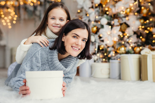 Brunette smiling female adult and her beautiful adorable small kid in white sweater being in living room, hold present, going to congratulate father and husband with coming New Year or Christmas