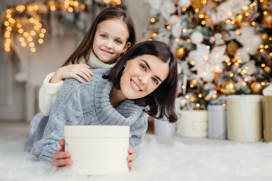 Children, family and celebration concept. Adorable female in knitted sweater holds white present box and small kid stands behind her back, have good mood before New Year`s Eve. Waiting miracle