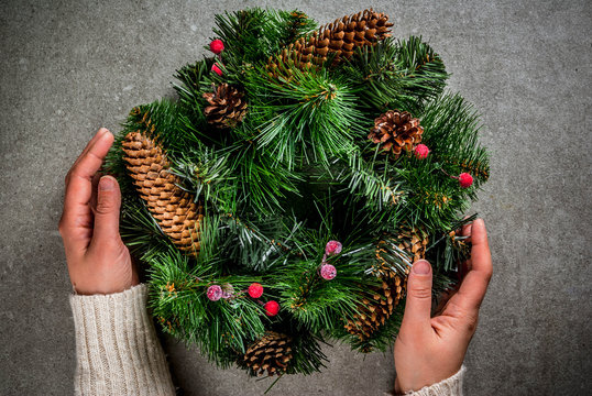 Preparation for xmas holidays. Woman decorating christmas green wreath with pine cones and red winter berries, on gray stone wall background, top view copy space, female hands in picture