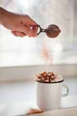 Cup of hot cocoa with marshmallows on white window sill, woman hand, vertical photo