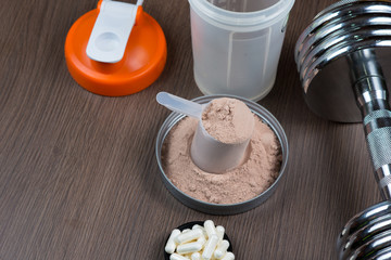 Whey protein in a measuring bucket with a metal dumbbell and shaker on a wooden background