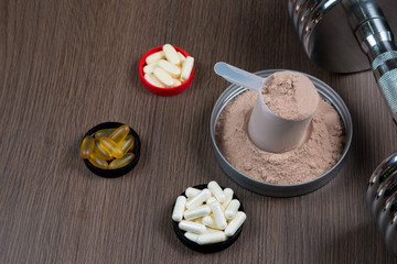 Whey protein in a measuring bucket with a metal dumbbell on a wooden background