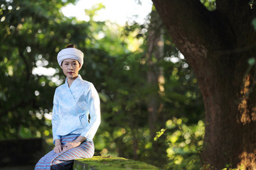 Fototapeta premium Portrait of thai young woman with traditional thai dress sit in thai temple with tree and sunrise