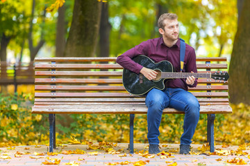 Young man playing acoustic guitar on bench in autumn park