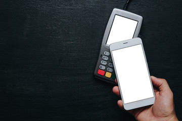 Top view of person using contactless payment with smartphone. Clear white mockup screen template of phone and pos terminal on dark wooden desk with side copy space