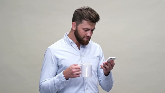 Shocked bearded man in shirt using smartphone while holding cup of tea over gray background