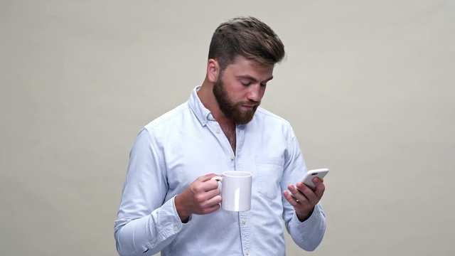 Tired bearded man in shirt using smartphone and yawns while holding cup of tea over gray background