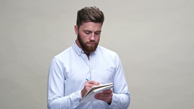 Concentrated bearded man in shirt listening someone and writing something in notebook over gray background
