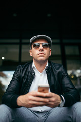 Fashionable retro dressed man with cap, suspenders and eyeglasses sitting on city street and looking at side. Low angle view.