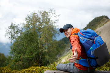 Naklejka na ściany i meble Image of sporty smiling man stretching out hand with backpack against backdrop of mountain