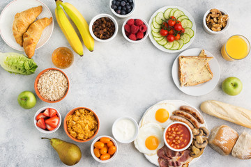 Full English breakfast frame, fried eggs, sausages, bacon and mushrooms with selection of fruits and vegetables, breads and juice on grey white table, top view, copy space for text, selective focus