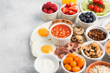Breakfast, fried eggs, sausages, bacon and mushrooms with selection of fruits and vegetables, orange juice on the grey white table, copy space for text, selective focus