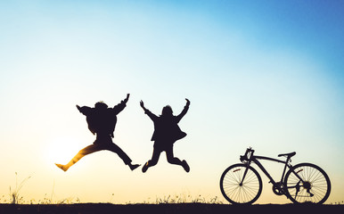 two people silhouetted against a sunset, jumping in the air with their arms raised. A bicycle is parked on the ground to the right.