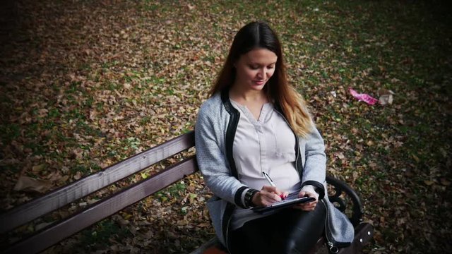 Beautiful young woman is writing a diary outdoors in the park, student studying on a bench in the park, Student girl preparing for the exam on the nature
