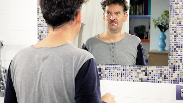 Man washing teeth in bathroom in pajamas in front of mirror unhappy about yellow teeth