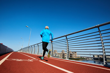 Rear view of active man in sportswear running down racetrack along railings of city bridge