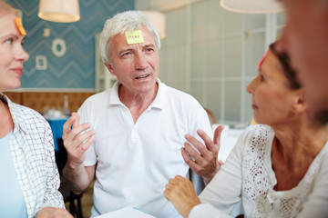 Grey-haired man explaining meaning of word from paper sticker on forehead to his friends