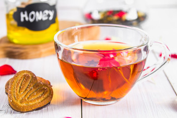 A cup of tea, biscuits, in the background of a can of honey and a pot of black herbal floral tea on a white wooden table.