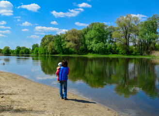 The young woman holds the child on hands and walks along the river in sunny spring day.