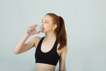 beautiful slender girl with red hair in sportswear on a light background with a bottle of water