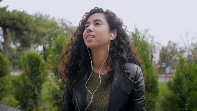 Pretty young hispanic girl with black curly long hair listening music in headphones on a deserted autumn street. Multi ethnic girl enjoying moment and singing. Slow motion.