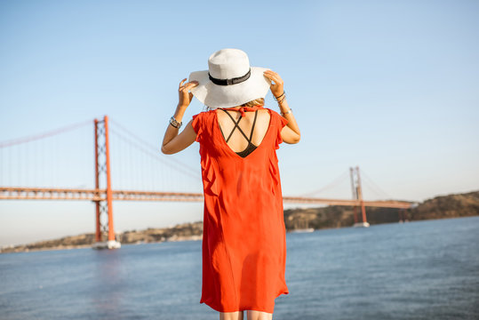Woman in red dress enjoying landscape view on the famous iron bridge standing back on the riverside in Lisbon city, Portugal