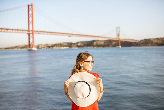 Portrait of a woman in red dress with sun hat standing on the riverside with beautiful iron bridge on the background in Lisbon city, Porugal