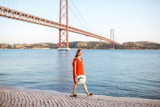Lifestyle portrait of a woman in red dress walking on the riverside with beautiful iron bridge on the background in Lisbon city, Porugal