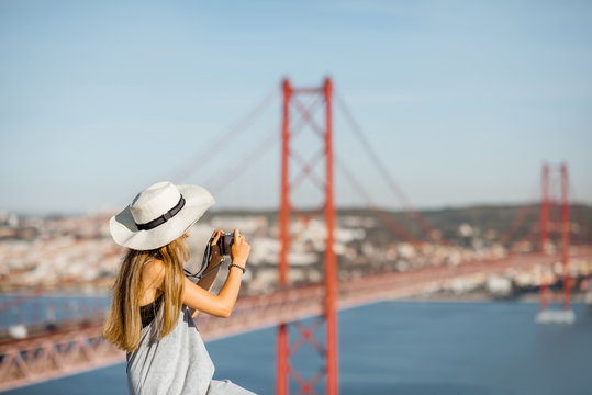 Portrait of a young woman tourist with photo camera sitting on the landscape view background with beautiful iron bridge in Lisbon city, Portugal
