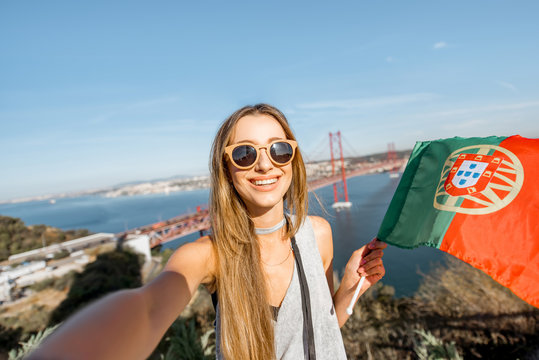 Young woman making selfie photo with portuguese flag on the beautiful landscape view background with bridge and river in Lisbon, Portugal