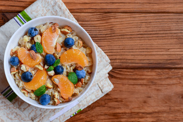 Light porridge with toppings. Oatmeal with juicy mandarins, blueberries, hazelnuts and mint in a white bowl and on a wooden background with copy space. Low calorie porridge recipe. Top view