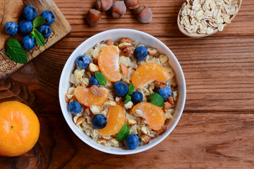 Easy porridge with fruits, berries and nuts. Classic oatmeal with juicy mandarins, blueberries, hazelnuts and mint in a white bowl and on a wooden table. Healthy porridge breakfast. Top view