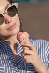 Young, beautiful brunette woman eating pink ice cream in a waffle horn. She wears glasses with dark glasses, and a shirt in a white-blue strip