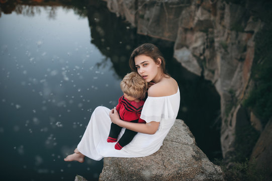 Young model-looking woman with a sad face sitting on the rocks near the river with a doll on her lap. A teenage girl imagines that she is a mother.