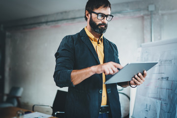 Bearded young coworker working at lightful office.Man using contemporary mobile digital tablet.Horizontal.Blurred background.